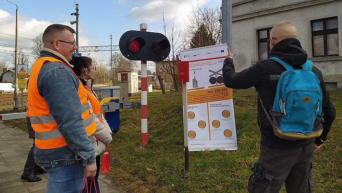 Sygnalizacja świetlna na przejeździe, obok banner żółtej naklejki, 2 instruktorów tłumaczy przechodniowi zasadę działania.