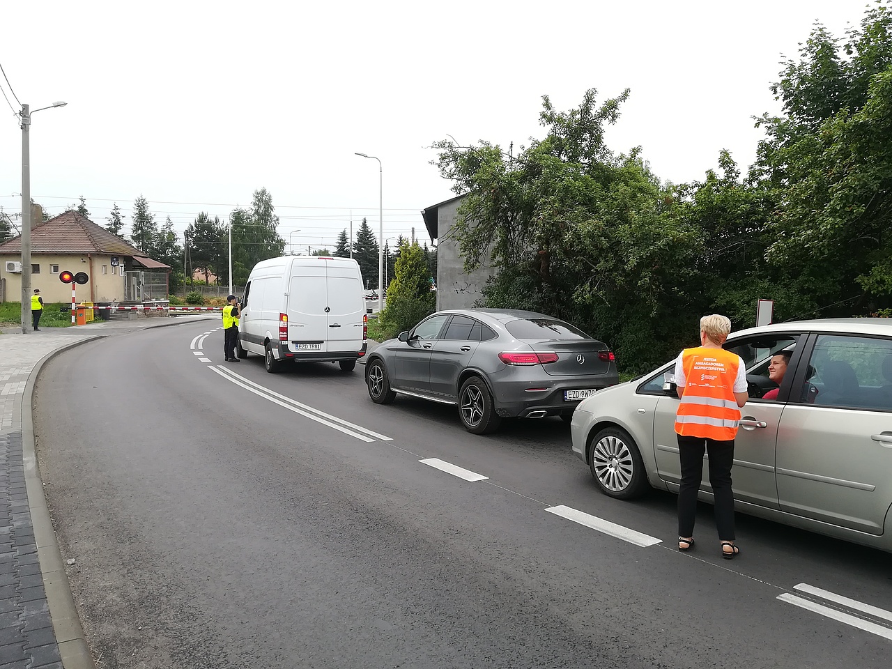 Przedstawiciel kampanii wręcza kierowcy w samochodzie materiały informacyjne. Samochody stoją przed zamkniętym przejazdem.