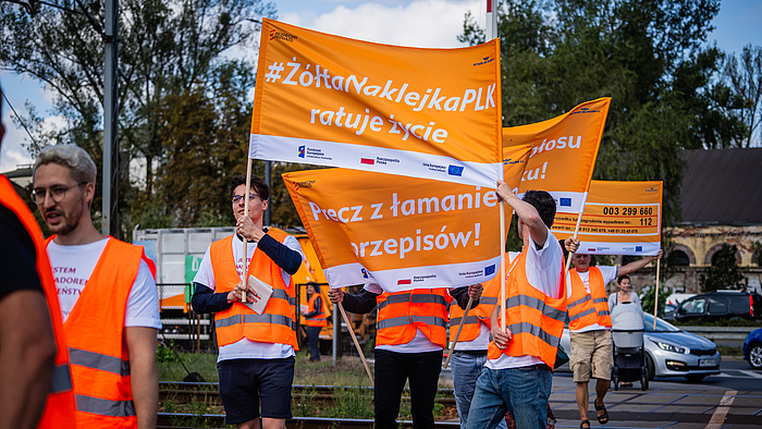Osoby w pomarańczowych kamizelkach trzymające transparenty żółtej naklejki PLK