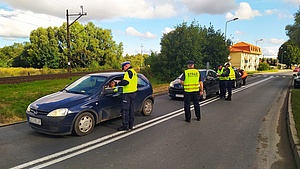 Policjanci z sokistami wręczają ulotki kierowcom w autach przed przejazdem kolejowo-drogowym.