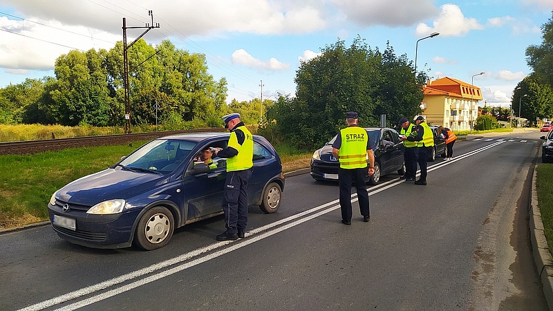 Policjanci z sokistami wręczają ulotki kierowcom w autach przed przejazdem kolejowo-drogowym.