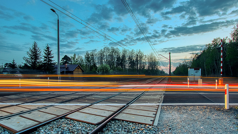 Przejazd kolejowy w wieczornych kolorach. Przez przejazd przebiegają poziome czerwone linie - efekt długiego naświetlania zdjęcia.