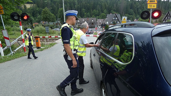 Przedstawiciel kampanii w asyście policjanta wręcza ulotkę kierowcy w samochodzie przed zamkniętym przejazdem.