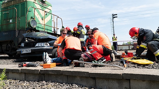 Grupa ratowników udziela pomocy poszkodowanemu kierowcy, w tle lokomotywa i wrak samochodu.