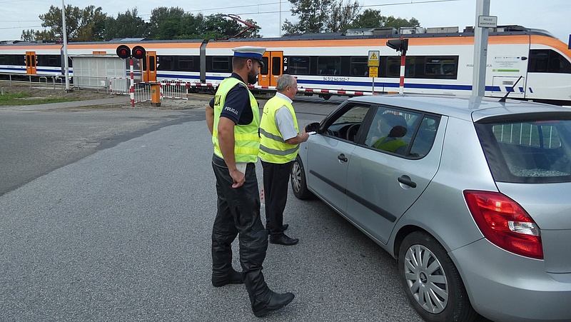 Przedstawiciel kampanii w asyście policjanta wręcza ulotkę kierowcy samochodu przed przejazdem. Na torach przejeżdżający pociąg