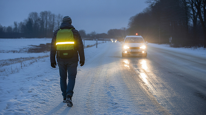 Mężczyzna idzie po oblodzonej drodze wieczorem, na plecaku ma odblask, na drodze samochód