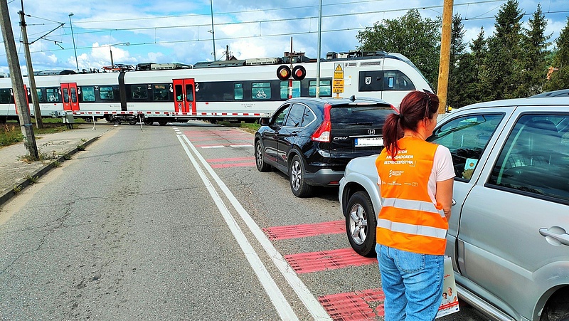 Pociąg na torach za zamkniętym szlabanem, przedstawicielka kampanii obok oczekującego samochodu przed przejazdem