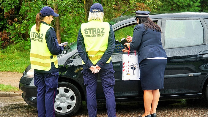 Funkcjonariuszki SOK oraz przedstawicielka kampanii w mundurze kolejowym wręcza materiały informacyjne kierowcy w samochodzie.