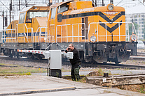 Moment korzystania z żółtej naklejki PLK, mężczyzna z telefonem przy szlabanie, w tle lokomotywa.