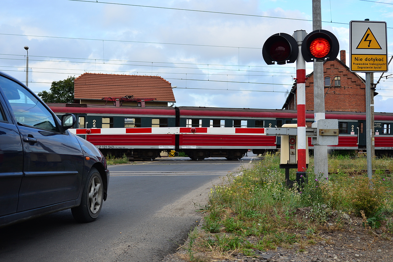 Auto stojące przed zamkniętą rogatką na przejeździe kolejowo-drogowym. W tle przejeżdżający pociąg.