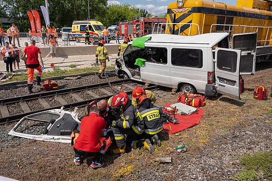 Grupa ratowników udziela pomocy poszkodowanemu kierowcy, w tle lokomotywa i wrak samochodu.
