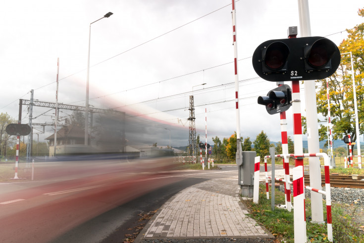 Przejazd kolejowy z sygnalizacją świetlną, z lewej strony rozmyty poruszający się pojazd w ruchu.
